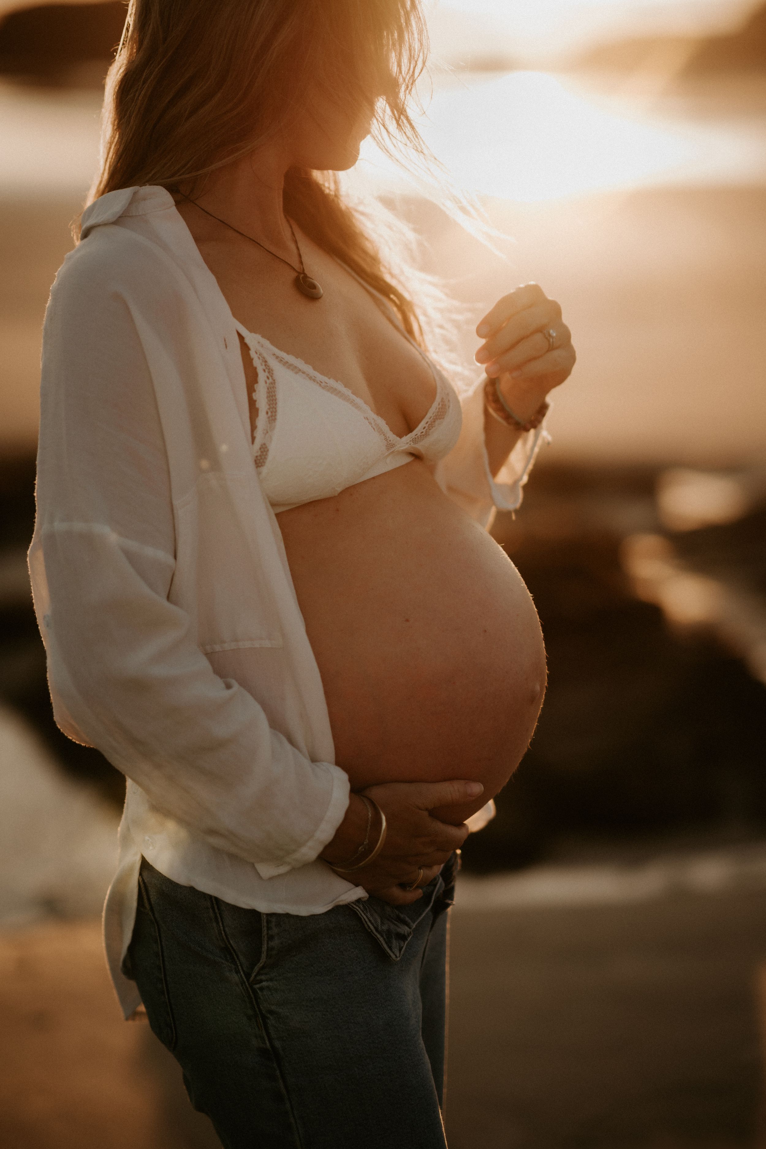 Pregnant woman in white shirt and jeans cradling baby bump at golden sunset on beach.