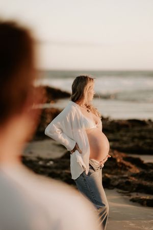 Pregnant woman in white shirt and jeans poses on rocky beach at sunset during maternity photoshoot.