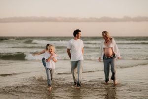 Pregnant woman and man walk on beach with young child splashing in waves at golden hour.