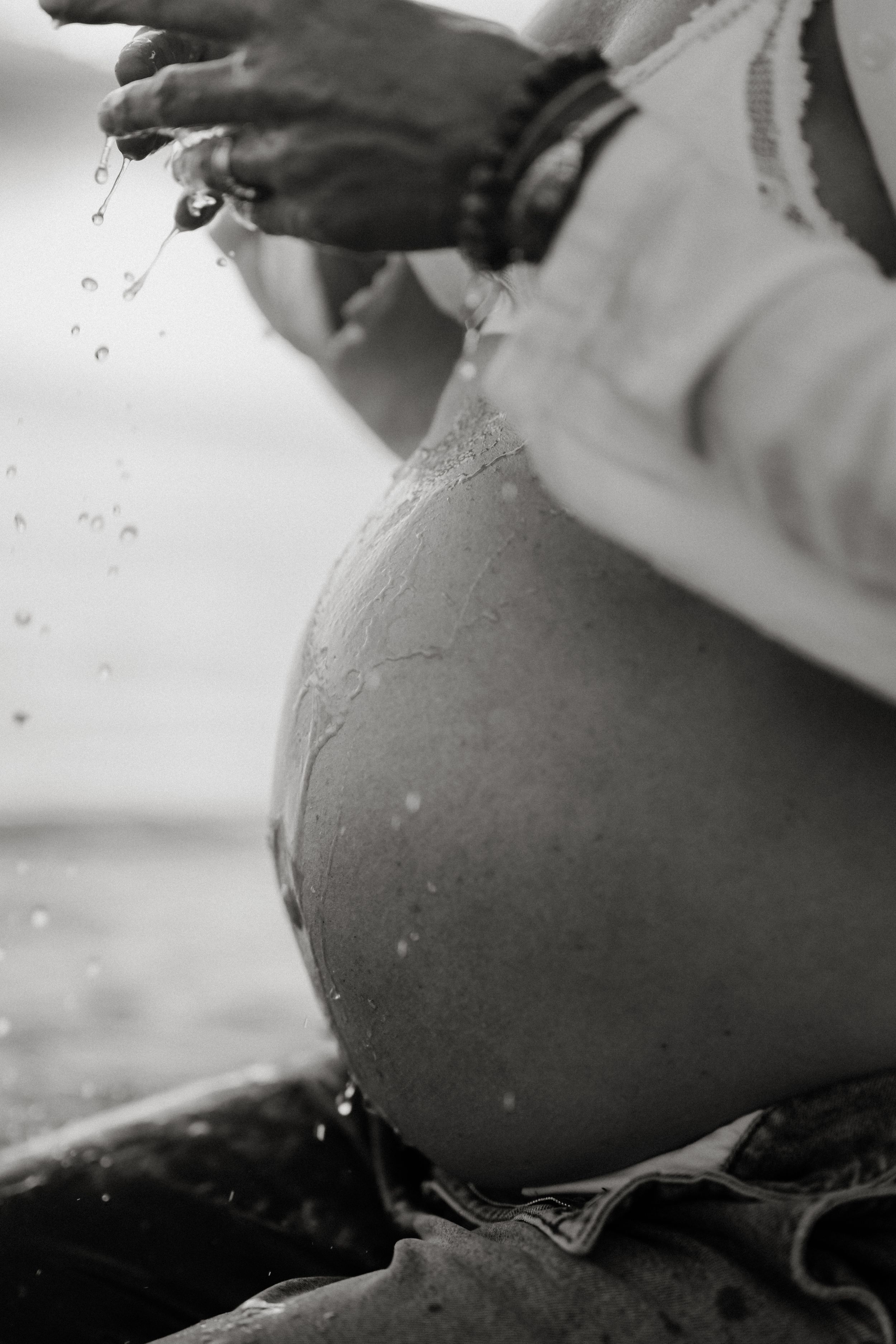 Black and white photo of a pregnant woman's bare belly with water droplets splashing from her hands outdoors.