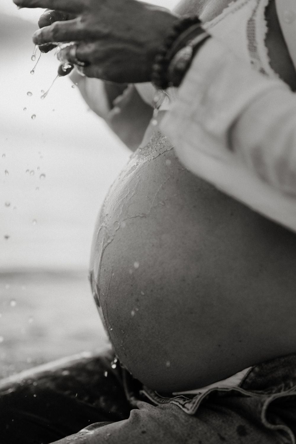 Black and white photo of a pregnant woman's bare belly with water droplets splashing from her hands outdoors.
