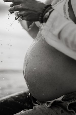 Black and white photo of a pregnant woman's bare belly with water droplets splashing from her hands outdoors.