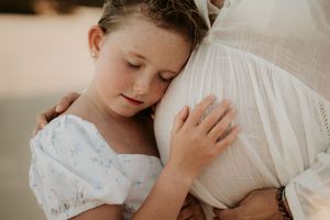 Young girl with freckles hugging a pregnant woman in a white dress outdoors in soft natural light.