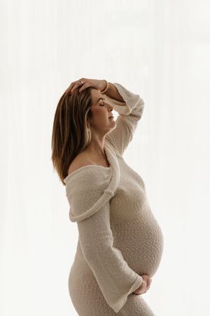 Pregnant woman in cream knit dress cradling her baby bump, posing gracefully against a soft white background.