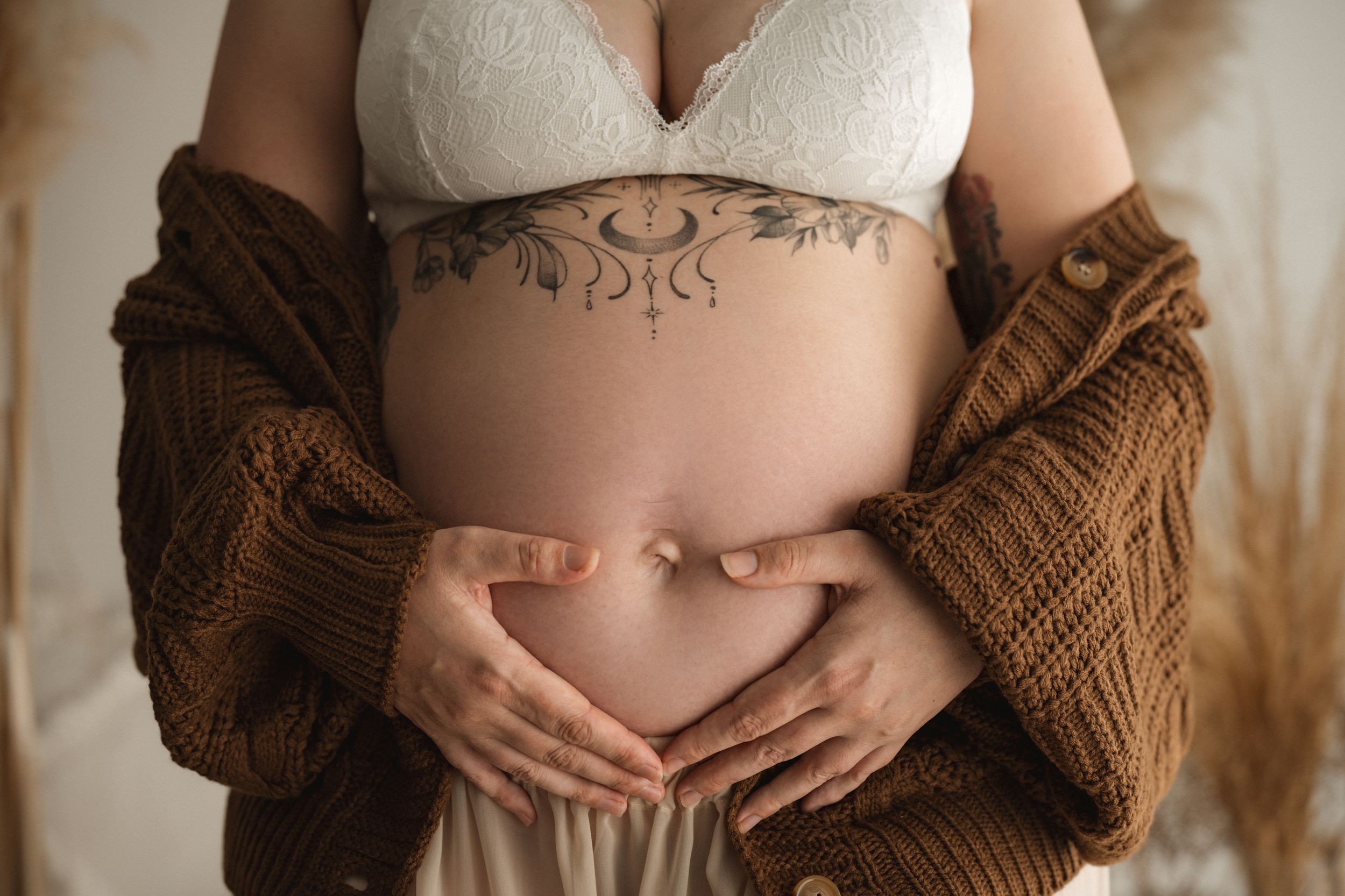 Pregnant woman cradling baby bump, wearing white lace bra, brown knit cardigan, with floral tattoo on abdomen.