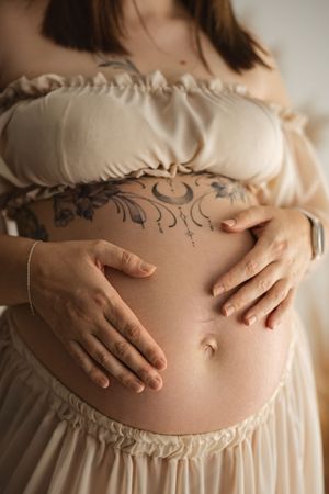 Pregnant woman in beige dress cradling tattooed baby bump with both hands, floral tattoo visible on abdomen.