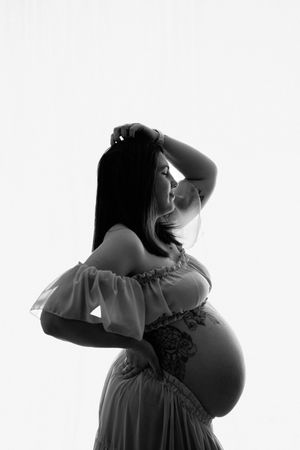 Black and white maternity photo of pregnant woman in lace gown, elegantly posing with hand in hair.