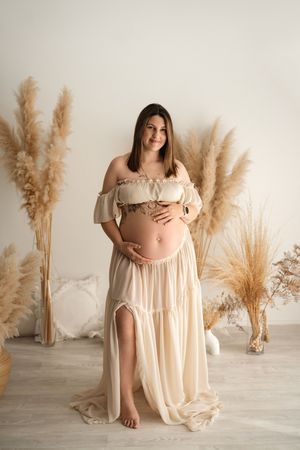 Pregnant woman in beige off-shoulder maternity gown holding baby bump, posing with pampas grass decor.