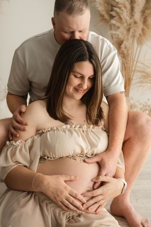 A couple shares a tender maternity photo, with the man embracing the pregnant woman from behind as both cradle her baby bump.