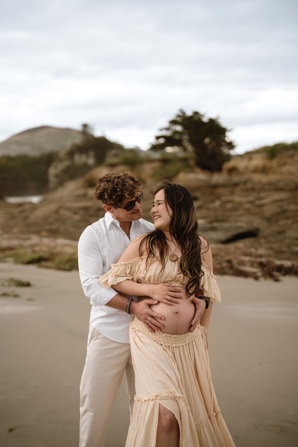 Couple embracing on beach during maternity photoshoot, woman in yellow dress, man in white, rocky coastline backdrop.