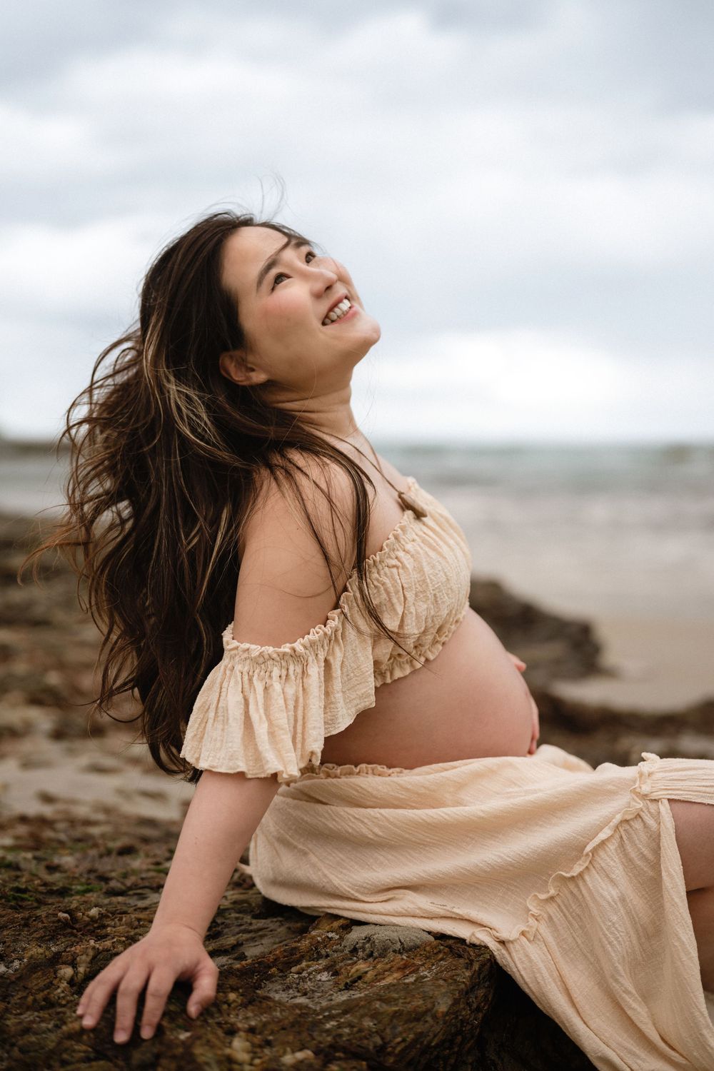 Smiling pregnant woman in cream off-shoulder dress sitting on rocks at beach, hair blowing in wind.
