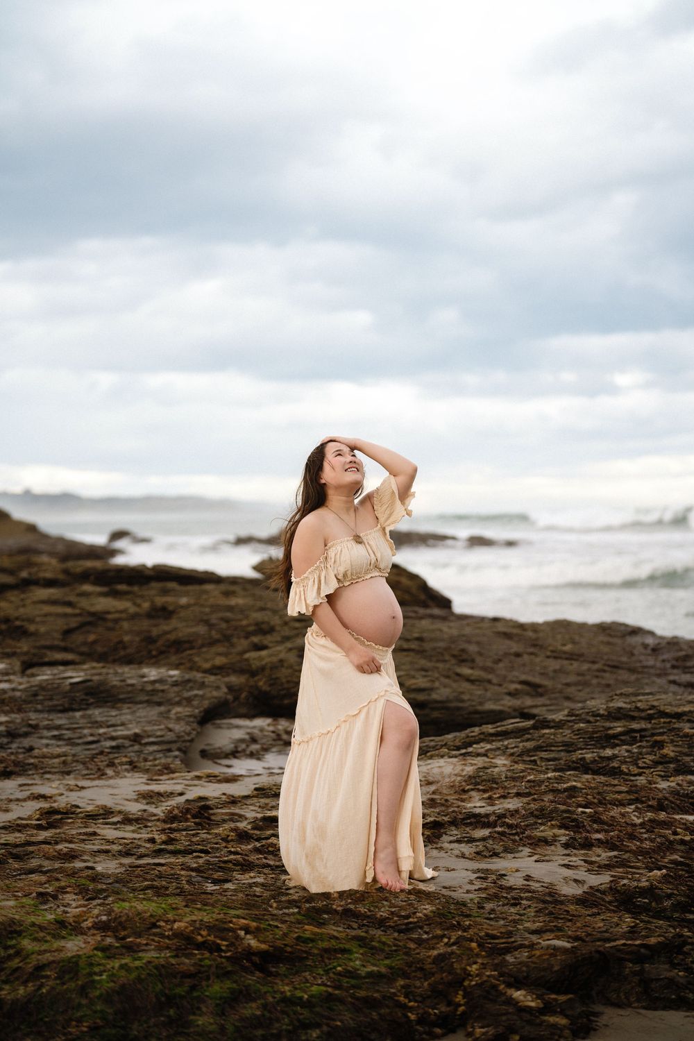Pregnant woman in flowing peach dress poses on rocky beach, cradling bump while gazing upward at cloudy sky.