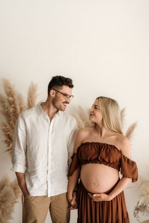 Couple sharing a tender moment during maternity photo shoot with pampas grass decor on neutral background.