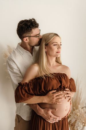 A man tenderly embraces a pregnant woman in a brown off-shoulder dress, kissing her head in a soft studio setting.
