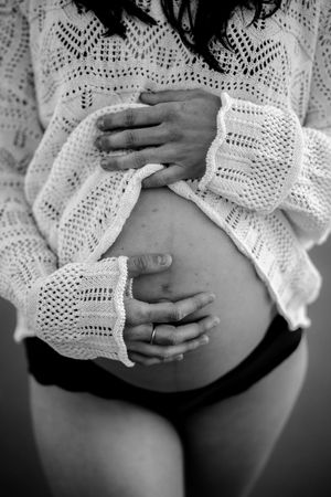Black and white photo of a pregnant woman cradling her bare baby bump, wearing a white knit sweater and black underwear.