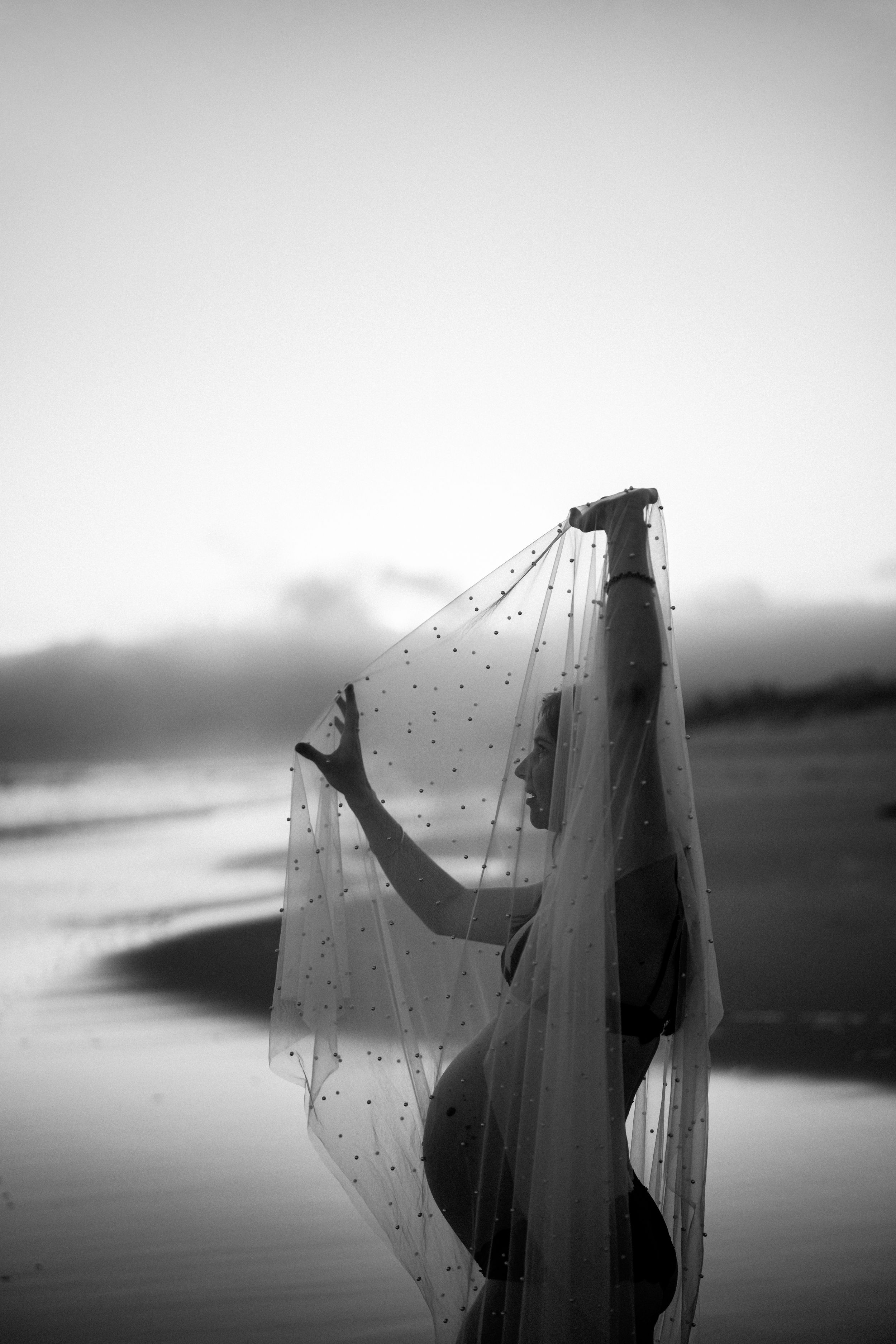 Black and white photo of a bride holding her dotted veil on a beach at sunset.