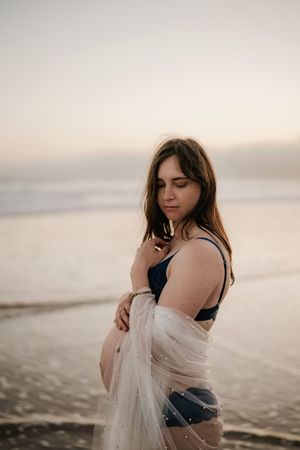 Pregnant woman in navy swimsuit and sheer wrap stands on beach at sunset, cradling her baby bump.