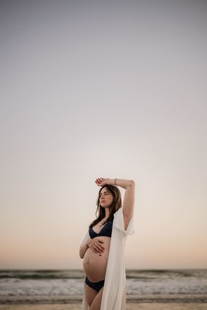Pregnant woman in black bikini and white kimono poses gracefully on beach at sunset, cradling her baby bump.