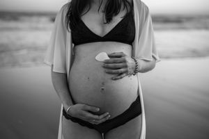 Black and white photo of a pregnant woman in bikini holding a seashell on her baby bump at the beach.