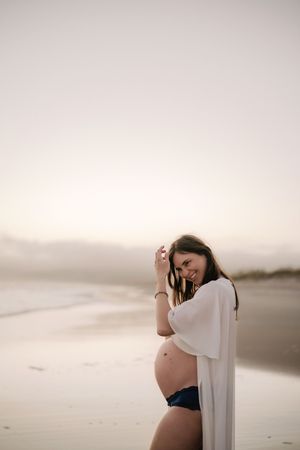 Smiling pregnant woman in white kimono and bikini posing on a serene beach at sunset with ocean waves in background.