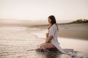 Pregnant woman in white shirt sitting peacefully on wet sand at golden hour beach sunset.
