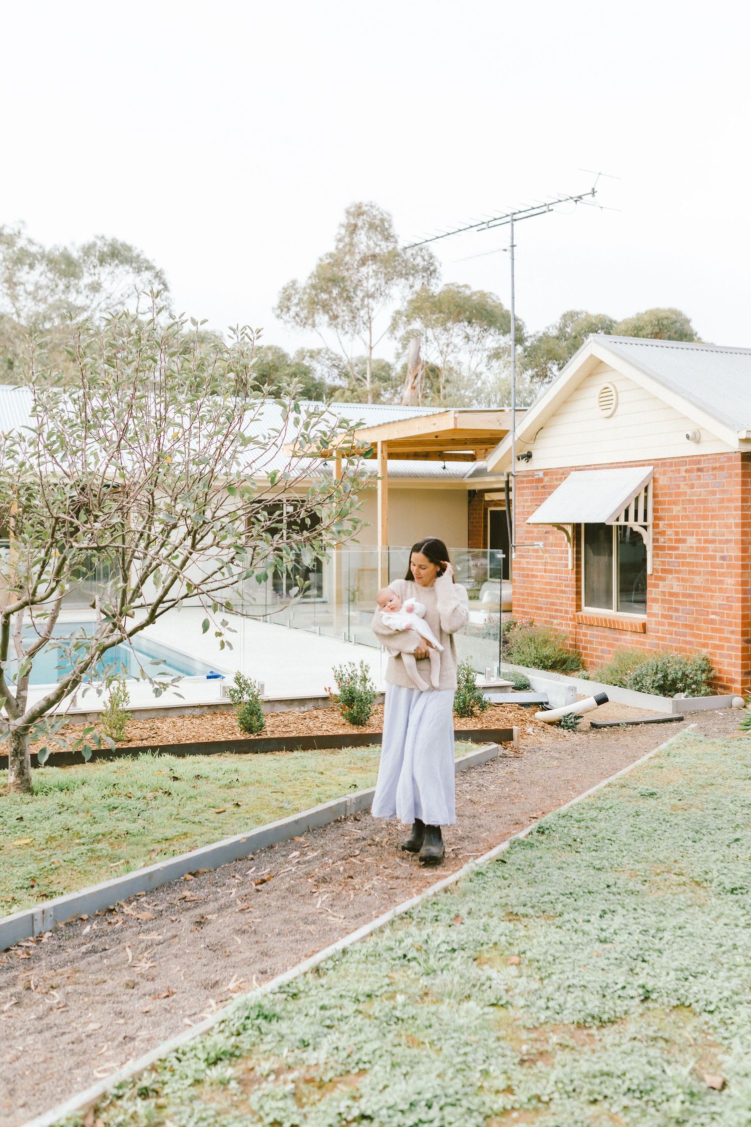 Woman in white dress holding newborn baby outside brick home with garden and pool in background.