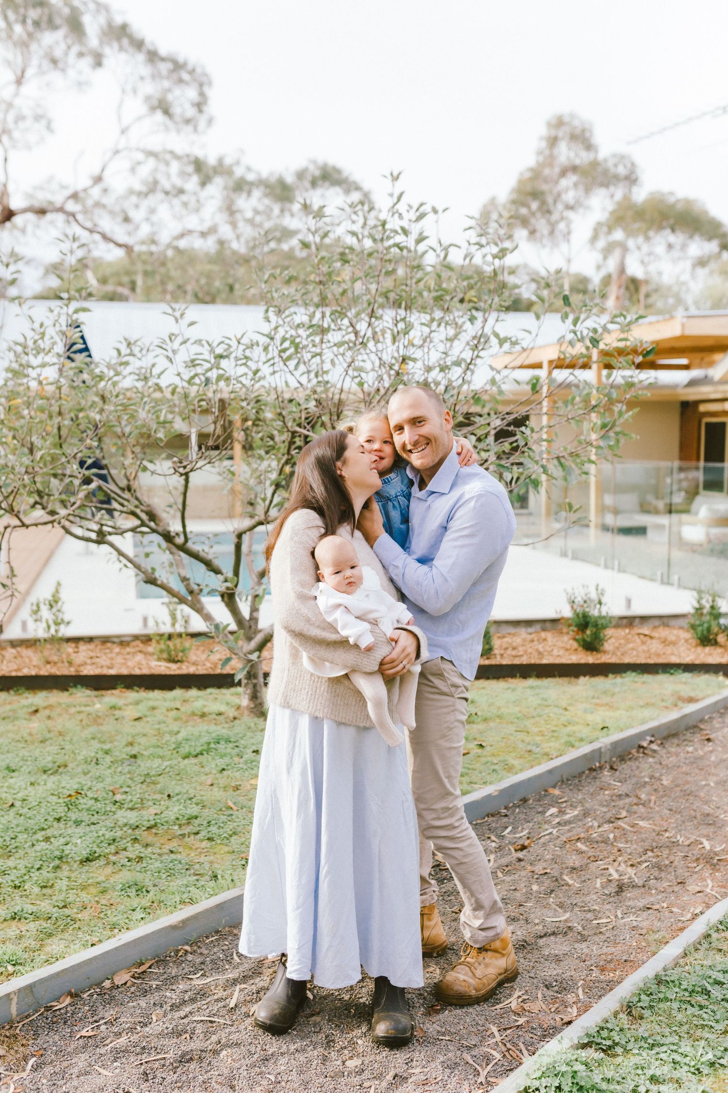 A happy couple shares a tender kiss outdoors while holding their newborn baby, surrounded by lush greenery.