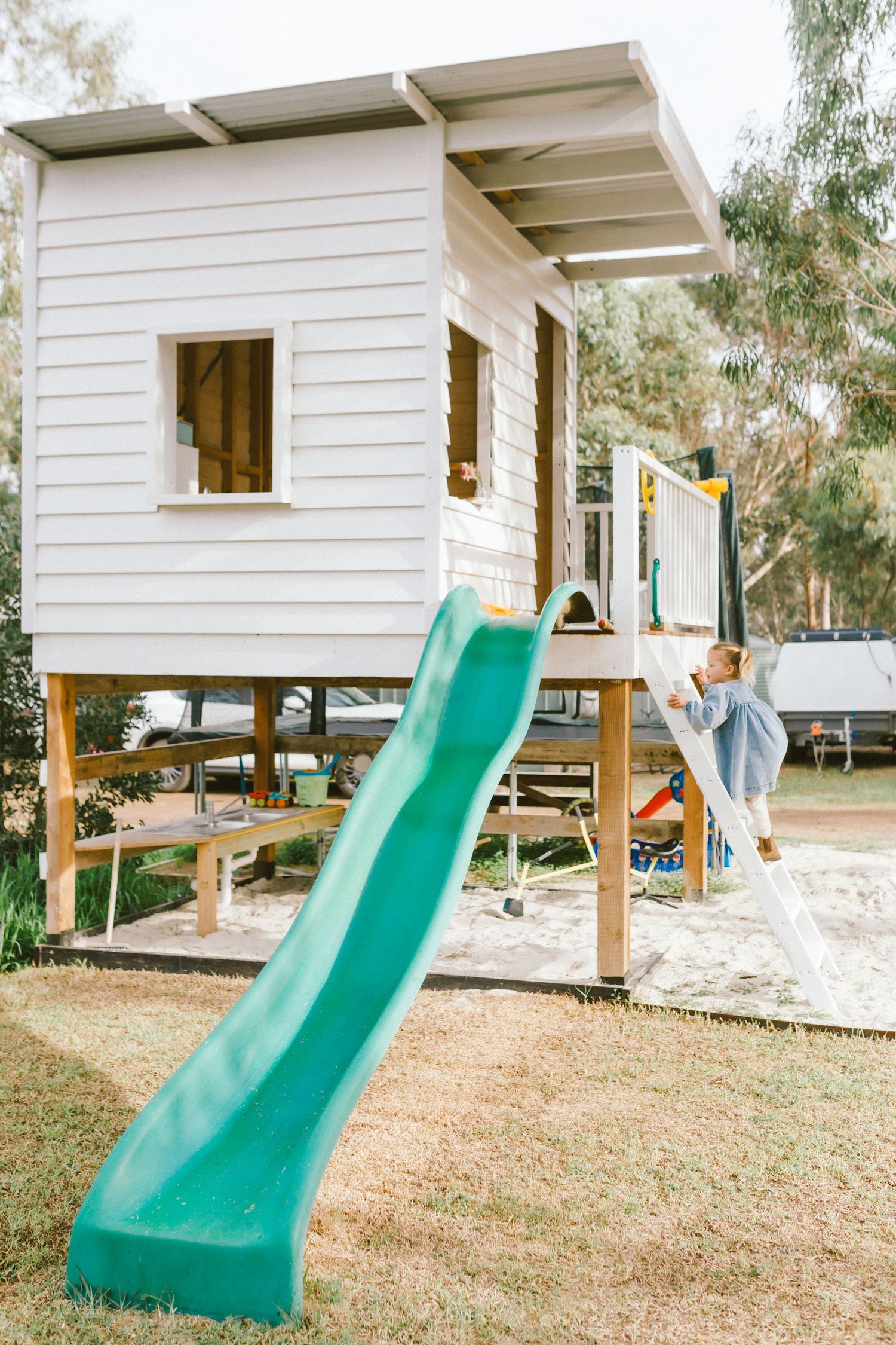 Child climbing ladder to elevated white playhouse with green slide in backyard playground on sunny day.