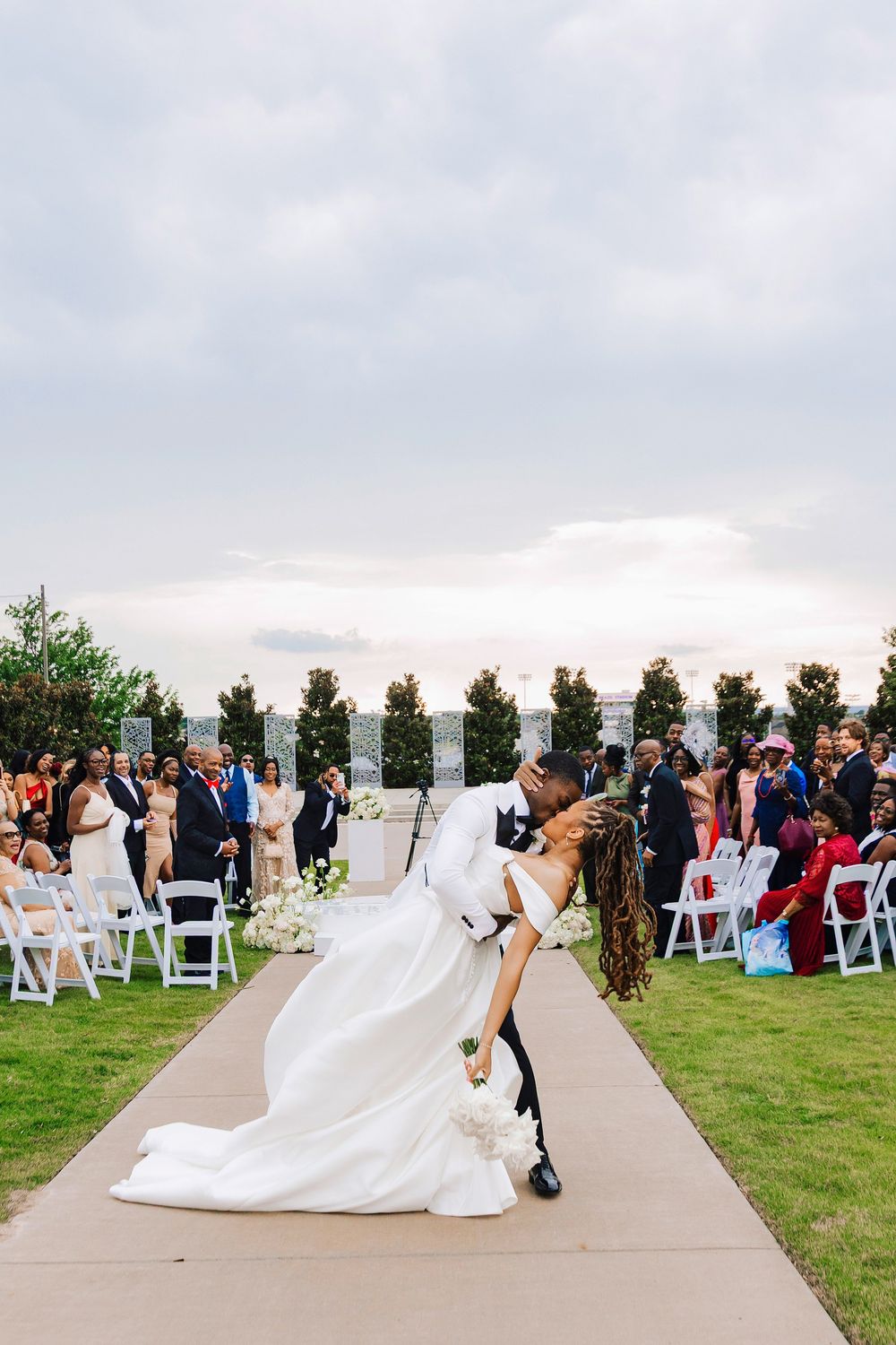 Groom dips bride for a romantic kiss at outdoor wedding ceremony with guests seated on white chairs.