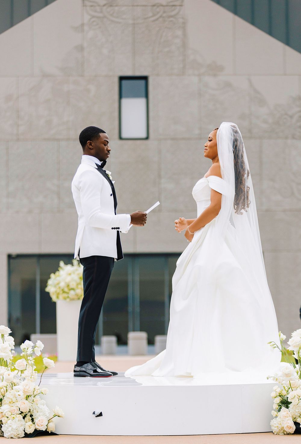 Bride and groom exchange vows outdoors on a white platform surrounded by white floral arrangements.