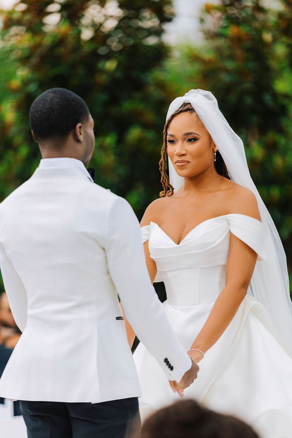 A bride in an off-shoulder white gown and veil holds hands with a groom in a white tuxedo during an outdoor wedding ceremony.