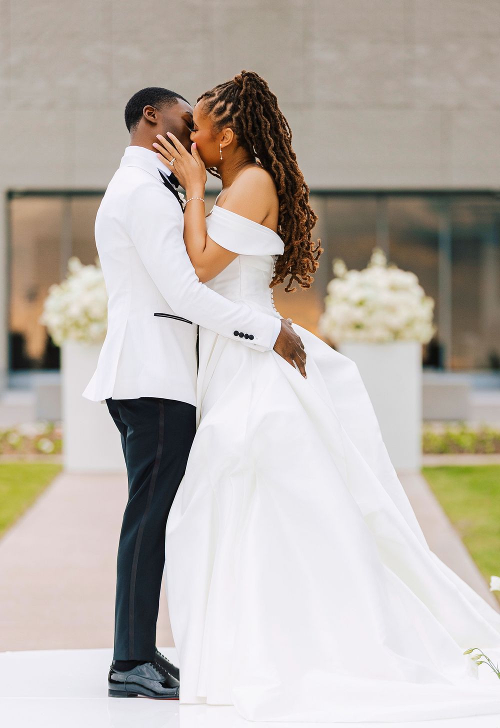 A bride and groom share a first kiss in elegant white wedding attire outdoors at a modern venue.