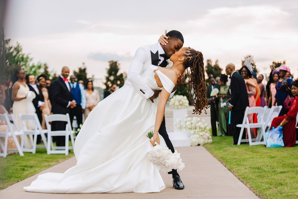 Groom dips bride for a romantic kiss at outdoor wedding ceremony with guests watching.