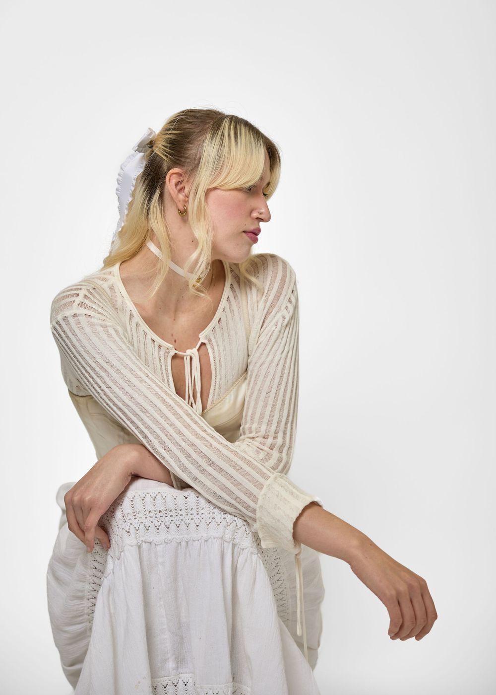 Blonde woman wearing a cream open-knit sweater with tie detail and white skirt, posing on white background.