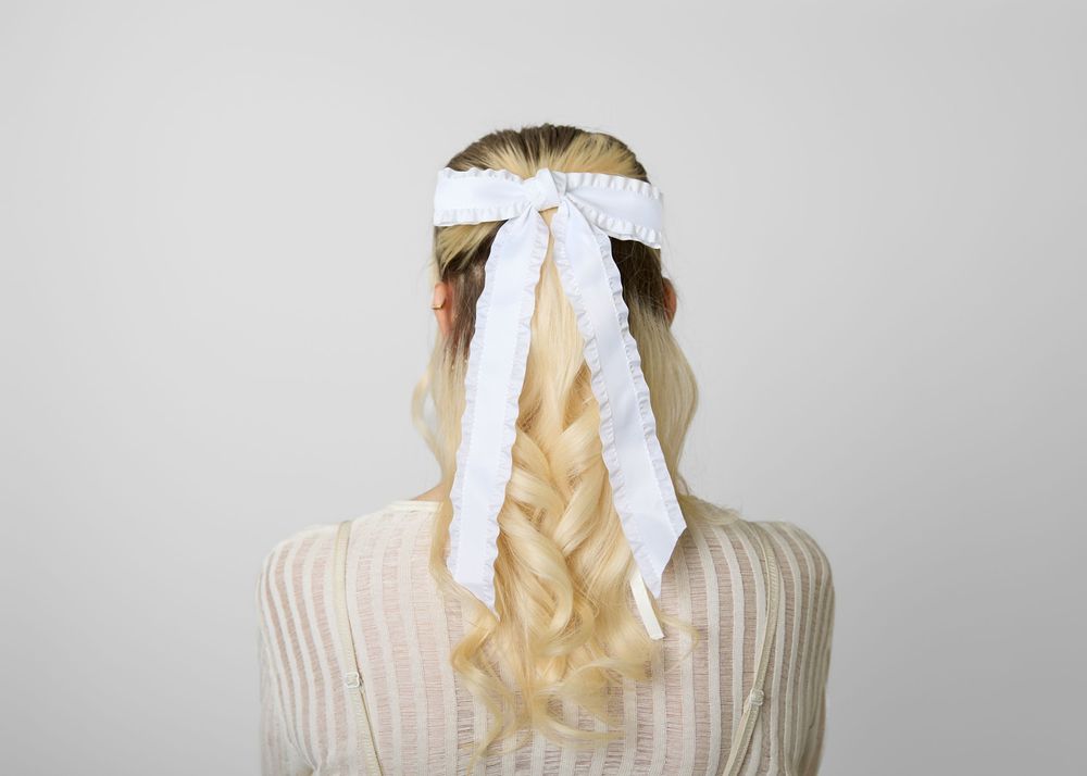 Blonde woman wearing a white satin hair bow with long ribbon tails in a half-up braided hairstyle.