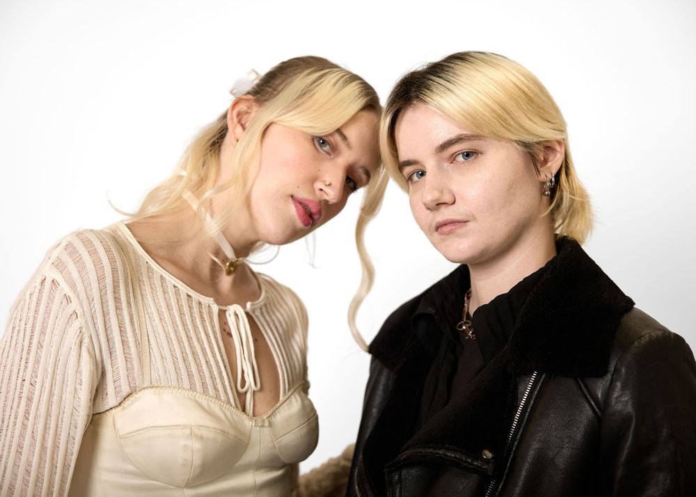 Two blonde women in cream and black outfits pose closely together in a studio portrait.