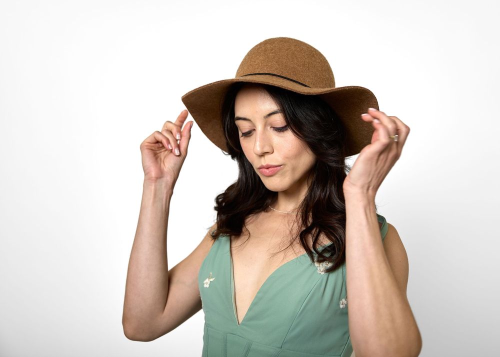 Brunette woman in green top adjusts wide-brim tan hat while looking down in studio.