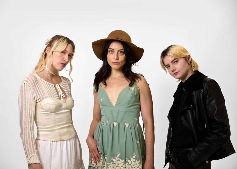 Three young women posing together, wearing a white lace dress, green floral embroidered dress, and black leather jacket.
