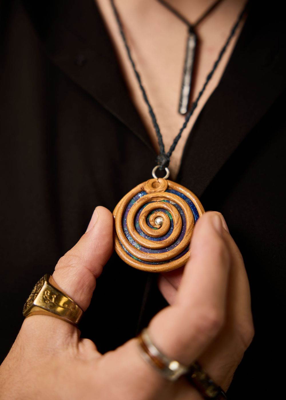 Hands holding a wooden spiral pendant necklace with blue inlay, worn over a black outfit.