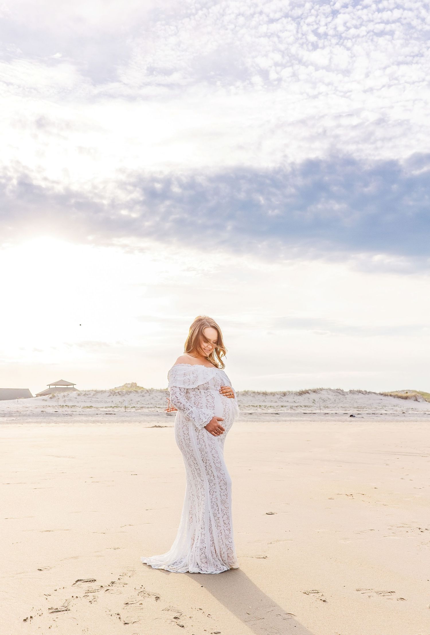 Pregnant woman in white lace off-shoulder gown on beach, cradling bump, soft clouds and sand dunes behind her.