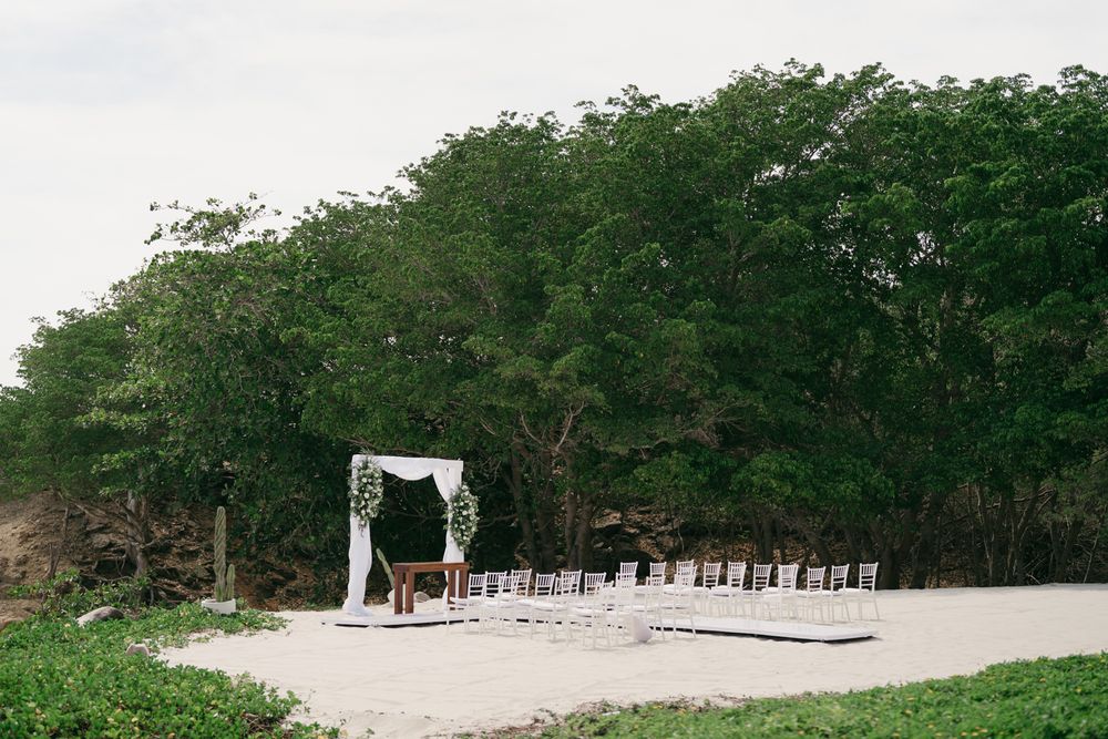 Outdoor beach wedding ceremony setup with white arch, chairs on sand, surrounded by lush green tropical trees.
