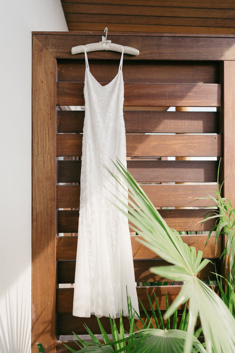 Elegant white beaded slip wedding dress hanging on wooden slat wall with tropical palm leaves.