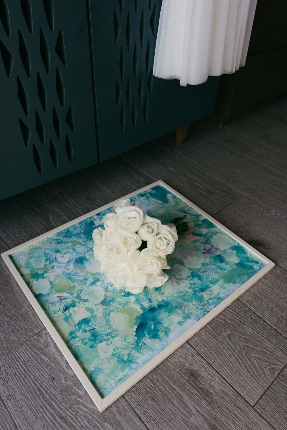 White rose bouquet resting on a teal watercolor painting on a gray wood floor near a teal cabinet.
