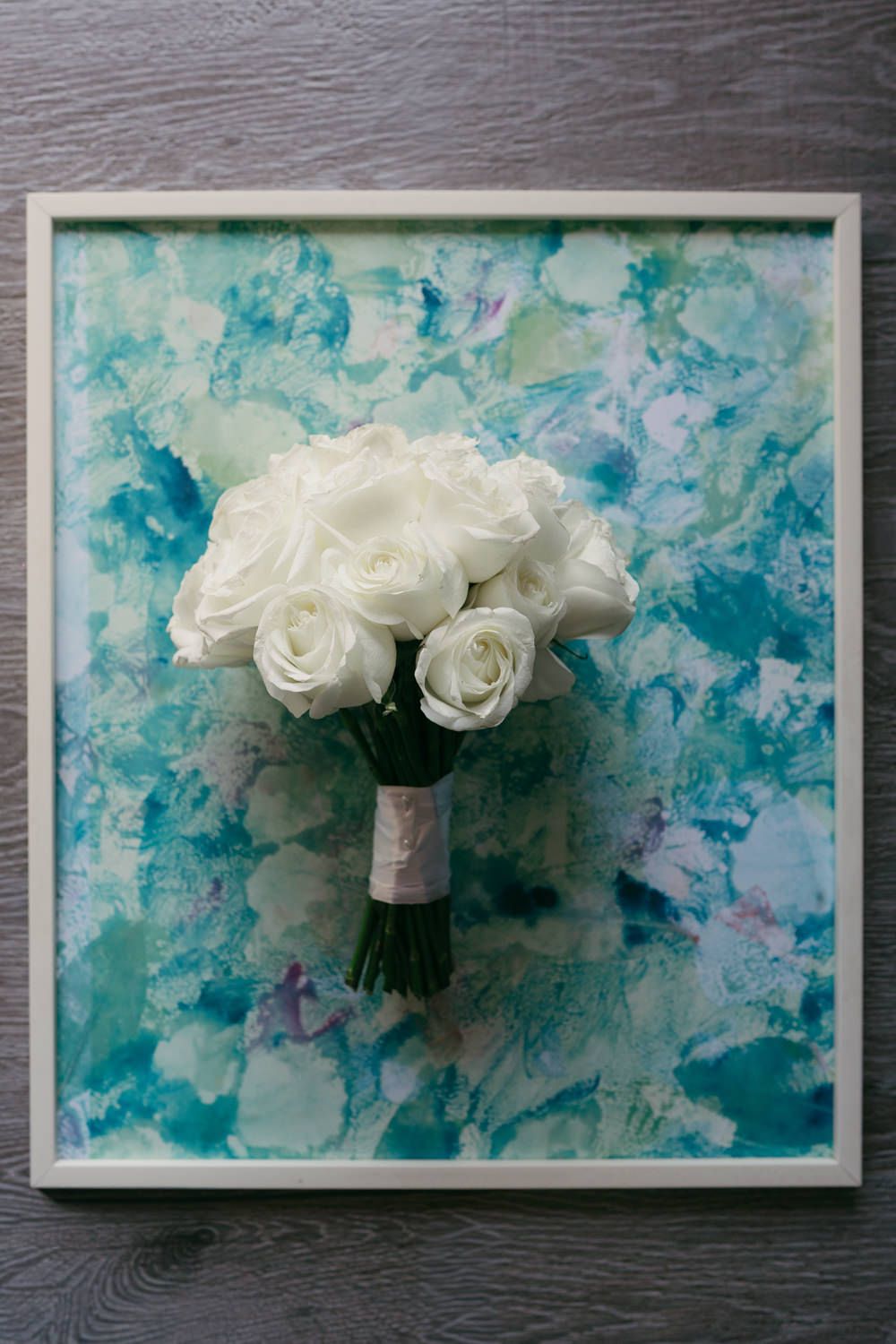 White rose bridal bouquet displayed against a framed watercolor teal and purple abstract painting.