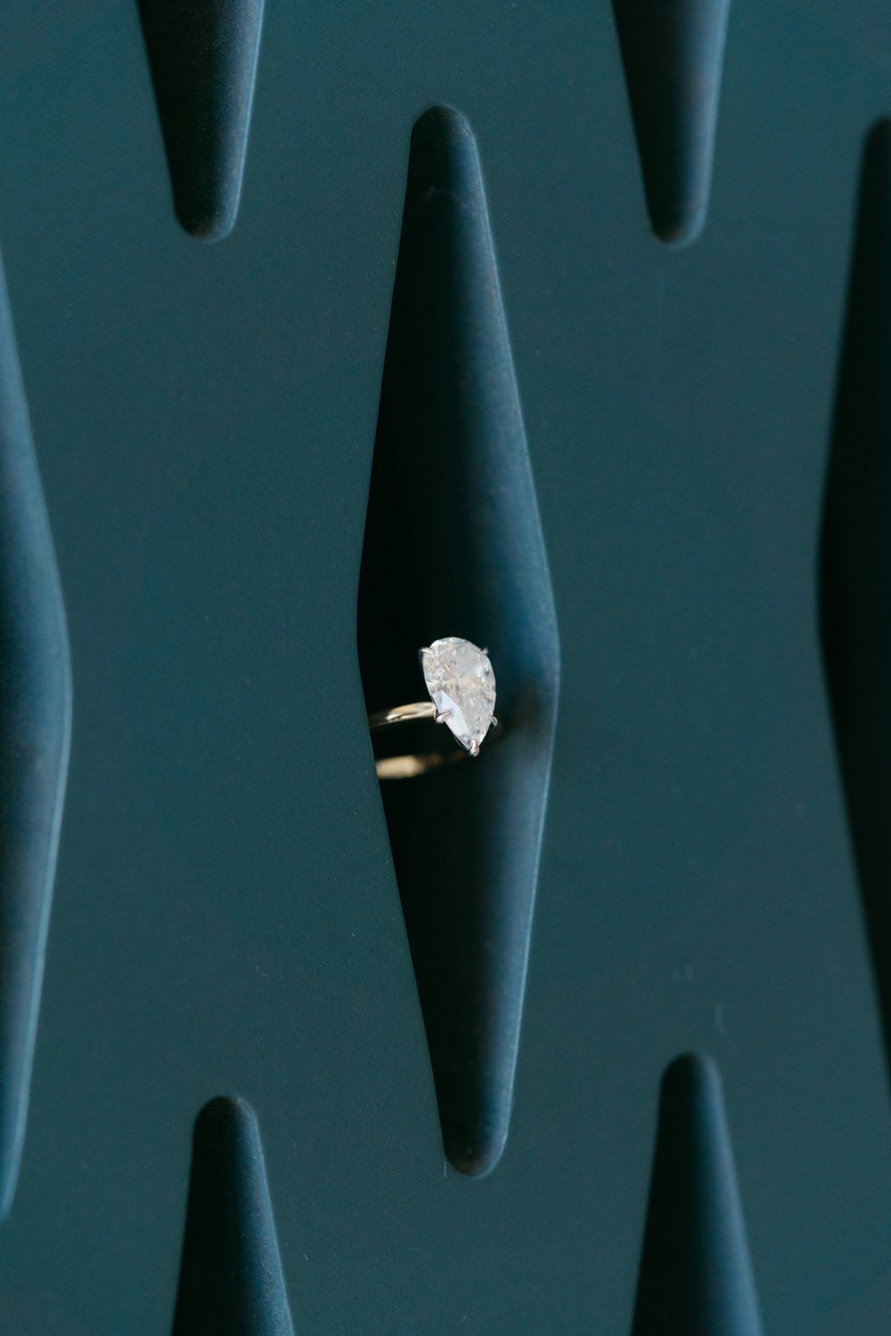 A pear-shaped diamond solitaire engagement ring displayed in a dark teal velvet ring box slot.