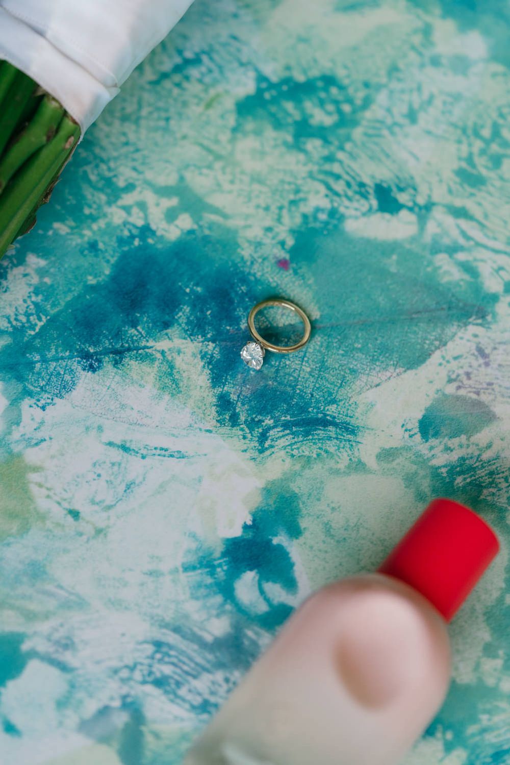 Two gold wedding rings, one with a diamond, displayed on a vibrant teal and white abstract painted surface.