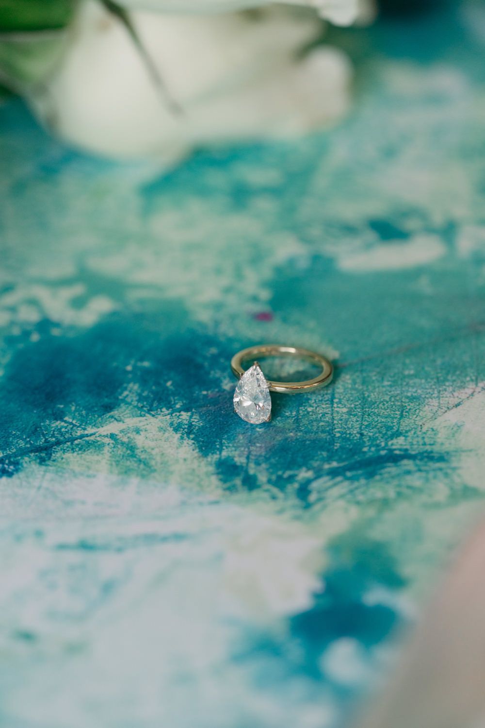 Pear-shaped diamond solitaire engagement ring displayed on a teal watercolor painted surface.