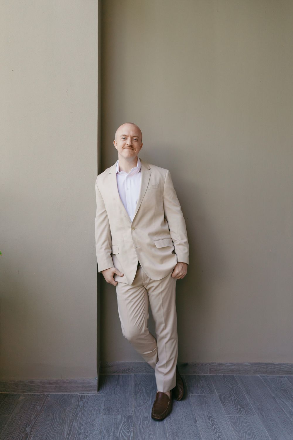 Bald groom in a beige suit leaning against a gray wall with one foot raised.