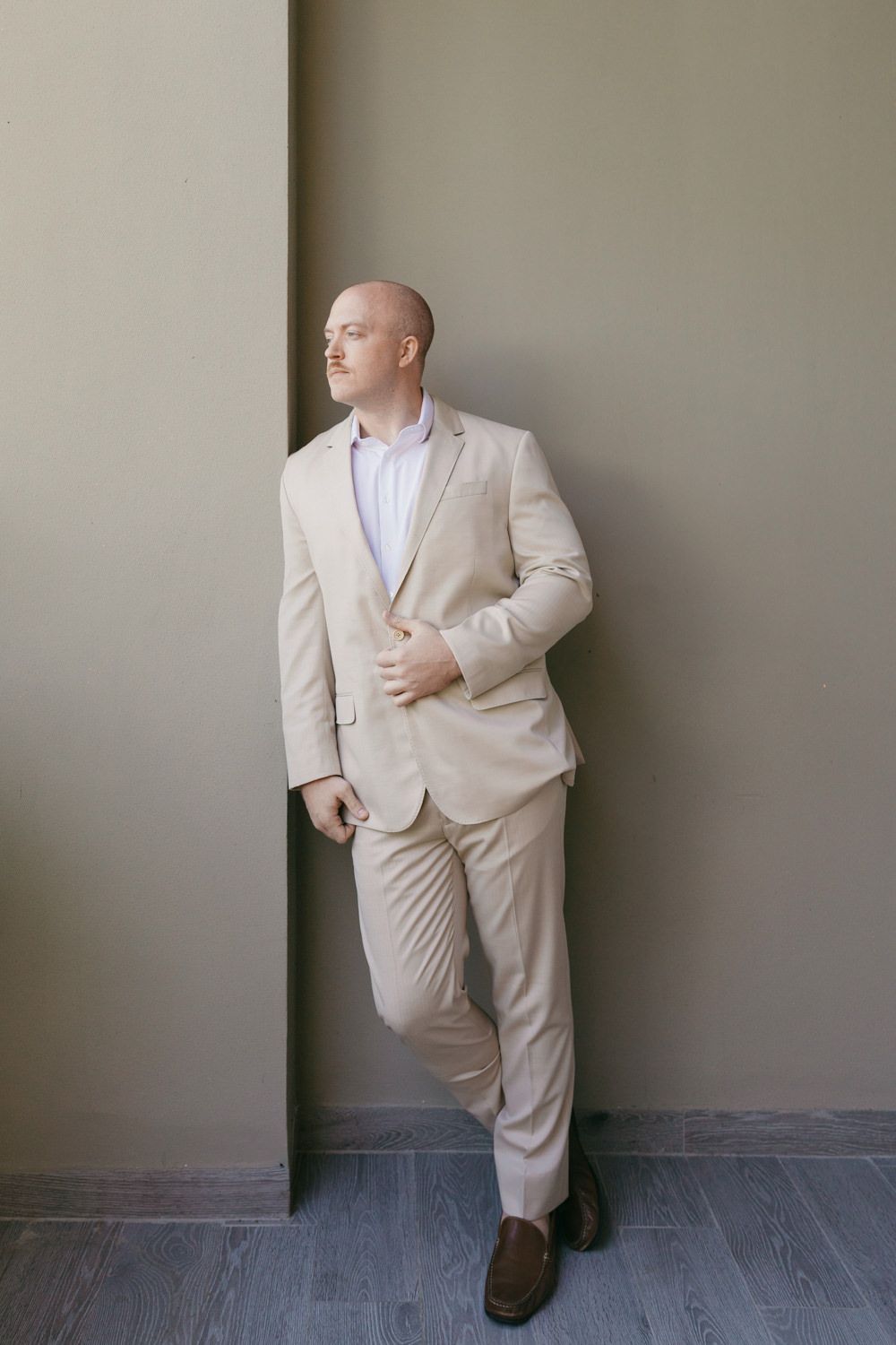 Bald groom in beige suit leaning against wall.
