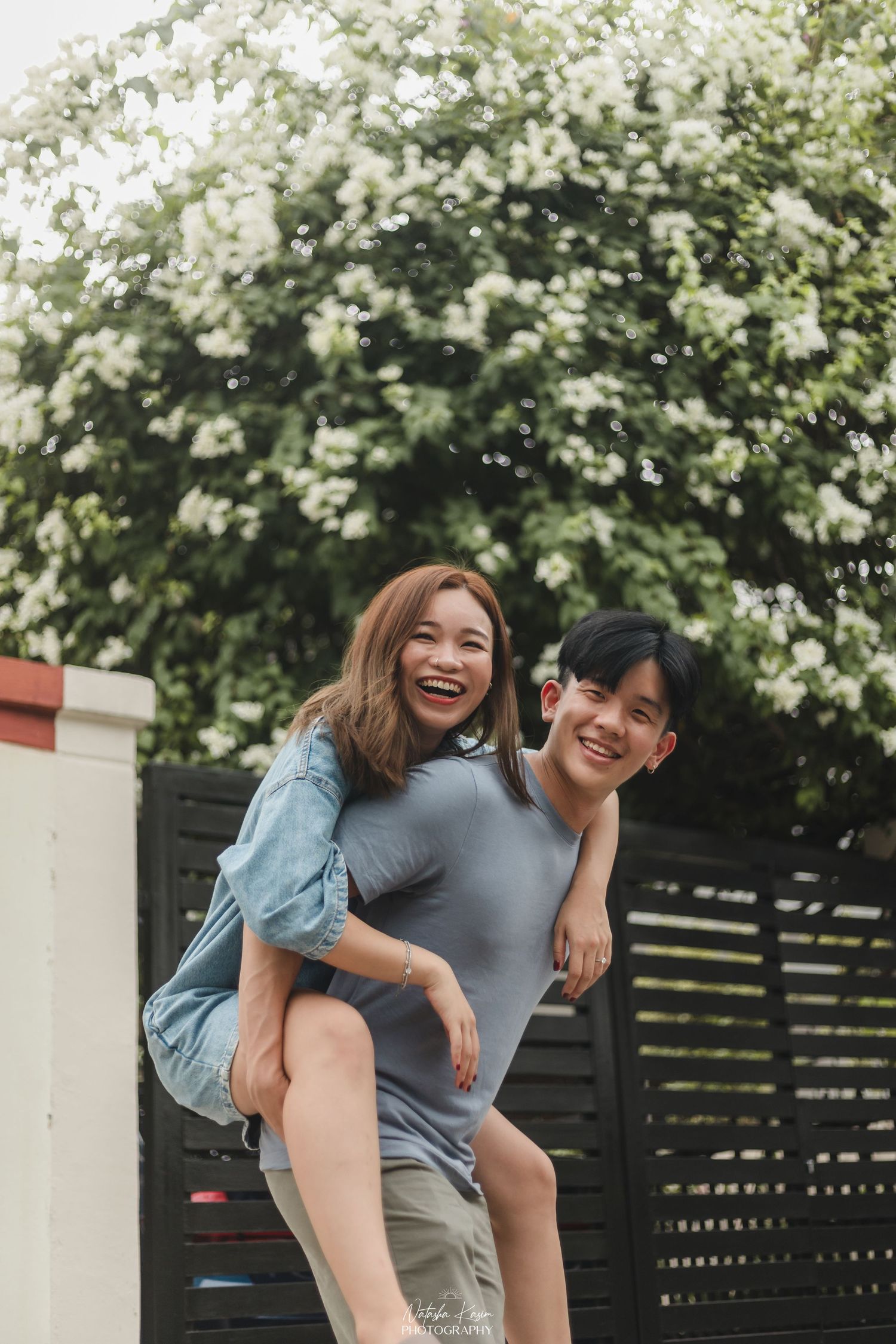 A joyful young Asian couple laughing outdoors, man giving woman a piggyback ride in front of white flowering bushes.